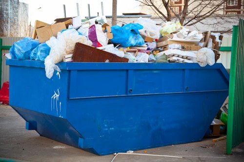 A large blue skip filled with a variety of waste materials including cardboard boxes, plastic bags, crumpled paper, and miscellaneous debris, situated on a paved surface in an outdoor urban area. The waste appears to be a mix of recyclable and non-recyclable items, with some cardboard boxes partially collapsed and plastic bags bulging with contents. The skip is positioned next to a green metal railing or fence, with a background of blurred trees and building structures, indicative of a typical commercial or residential waste collection site in Richmond upon Thames. Natural daylight illuminates the scene, highlighting the textures of the debris and the finish of the metal skip. This image reflects the type of waste managed by commercial waste removal services, such as those offered by Commercial Waste Removal Richmond upon Thames, and emphasizes the importance of proper rubbish disposal in the local area.