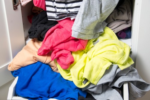A close-up of an open white laundry drawer filled with a mix of colourful clothing including a bright yellow garment, a red piece of fabric, a grey item, a blue garment, and various other clothes, all piled haphazardly in a domestic setting. The surrounding environment suggests a typical household space, possibly a bedroom or utility area, with the focus on the clothing clutter. The image captures the textures and different fabrics of the clothes, emphasizing their casual and used condition. This scene relates to waste management services provided by Commercial Waste Removal Richmond upon Thames, which handles the collection and disposal of laundry and clothing waste from household or commercial premises in the Richmond area while ensuring proper waste procedures are followed.