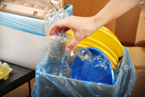 A close-up of a person's hand placing a clear plastic water bottle into a large blue recycling bag, which already contains several yellow and blue plastic buckets. The bag is set on a black surface in an indoor environment, with a white cardboard box and some yellow packaging material visible in the background. The scene appears to depict waste collection or recycling preparation, relevant to commercial waste removal services in Richmond upon Thames. The person's arm and hand are visible, and the plastic bottle is partially crushed, showing the typical transparency and smooth texture of PET bottles. The yellow buckets have a sturdy plastic finish, and the blue buckets appear to be slightly glossy. This detailed visual emphasizes the sorting and disposal of recyclable waste within a professional waste management context, aligning with services offered by Commercial Waste Removal Richmond upon Thames on their webpage about rubbish collection strategies in the local area.